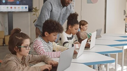 Medium slowmo of contemporary young male Afro American IT teacher helping 5 or 6 grade students working with laptops in class - Powered by Adobe