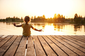Silhouette of a child sitting on the wooden pier and enjoying heat summer evening at the lake at sunset