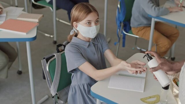 High-angle medium slowmo of 12-year-old school girl in face mask sitting at desk in classroom holding out her hands as unrecognizable male African-American teacher pouring some sanitizer on them