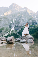 The bride and groom near the lake in the mountains. Newlyweds together against the backdrop of a mountain landscape. Romantic wedding couple in love standing on the stony shore. Scenic mountain view.