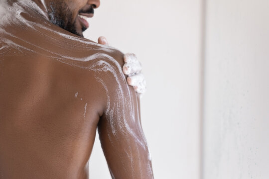 Muscular back of mixed race African bearded guy, taking shower, applying foamy gel or soap on smooth dark skin. Male morning bath routine, daily hygiene, bodycare concept. Cropped shot, close up