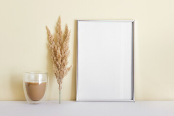 Mockup template with large silver A4 blank frame and coffee with dry yellow plant next to yellow wall.