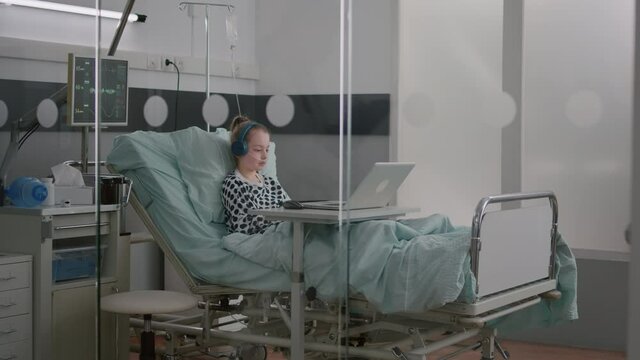 Alone sick girl patient with oxygen nasal tube relaxing in bed wearing headphones while watching entertainment movie on laptop computer during medical examination. Girl recovery after surgery