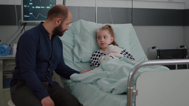 Sick girl child lying in bed discussing with worried father during diagnosis consultation in hospital ward. Hospitalized kid with oxygen nasal tube holding teedy bear in hands suffering flu infection