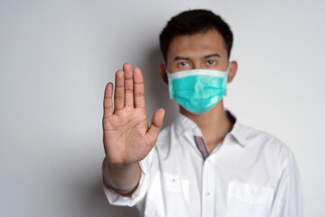 Portrait of young asian man wearing health mask with stop hand gesture in front of camera against white background. Symptoms of contracting corona virus concept. Novel Coronavirus (COVID-19)