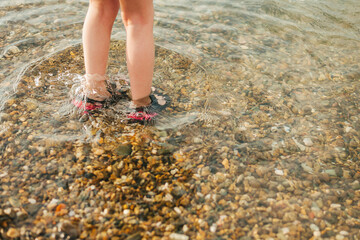 childrens bare feet in rubber sandals stand on a pebble beach in sea water summer