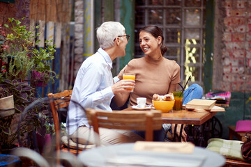elderly woman and young adult female having breakfast together, looking each other, smiling.