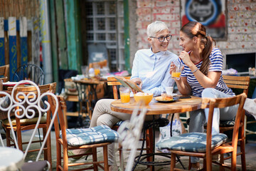 elderly woman showing part in opened book to young adult brunette female drinking orange juice on a...