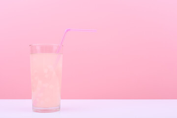 Close up of transparent glass with light pink fruit drink or lemonade with pieces of fruit and pink straw on white table against pink background with copy space. Trendy summer composition. 