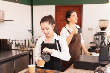 Asian barista women fill milk into takeaway hot coffee cup for customer while caucasian barista woman enjoy hot black coffee in cafe coffee shop