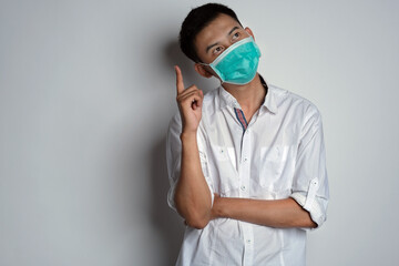 Attractive young asian man wearing health mask with thinking expression pose and his hand is pointing up, against white background. Corona virus photo concept. Novel Coronavirus (COVID-19)