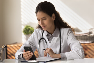 Young Caucasian female GP in white medical uniform sit at desk in hospital use modern cellphone...