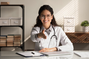 Portrait of young female doctor in white uniform stretch hand for handshake greet get acquainted with patient in clinic. Smiling woman GP excited to meet client in hospital. Good medicine concept.