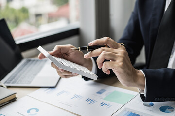 Naklejka premium A man with a pen pointing to a white calculator and documents are placed on the table, a businessman in a black suit is sitting to check information from documents, he is a company executive.