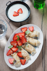 White oval plate with vareniki or curd dumplings, fresh strawberry and sour cream, vertical shot on a brown wooden background