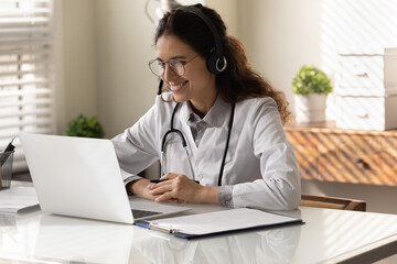 Smiling Caucasian female doctor in medical uniform headphones talk on video call on computer with...