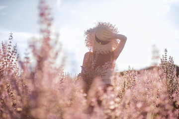 Young blonde woman in straw hat looks into the distance against the background of blooming field pink sage. Close-up portrait of beautiful girl holding bouquet flowers in hands. Agricultural texture