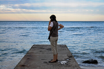 Unrecognizable Lonely Woman in Tracksuit is Standing with her Back and Talking on Phone Against the Background of Sea. The Concept of Waiting, Farewell to the Sea. Selective Focus. Life Style. 