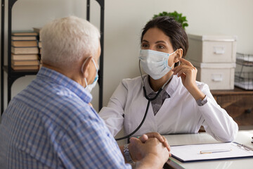 Young female doctor in facemask listen to old male patient heart chest with stethoscope at clinic meeting. Woman GP checkup examine mature man client with phonendoscope. Elderly healthcare concept.