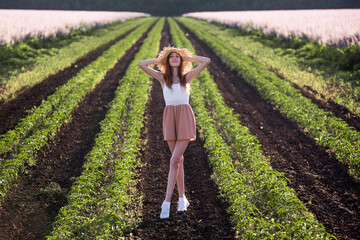 Young woman in white T-shirt, powdery shorts runs along green plowed field. Long hair develops at sunset, the girl has straw hat. Promote agricultural perspective, the texture of black soil ground