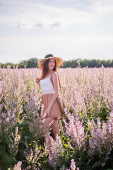 Young blonde woman in straw hat looks into the distance against the background of blooming field pink sage. Close-up portrait of beautiful girl holding bouquet flowers in hands. Agricultural texture