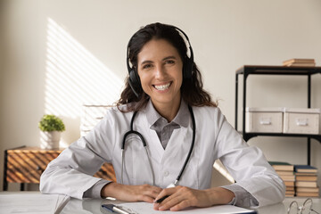Portrait of smiling young Caucasian female doctor in white uniform headphones have webcam digital...
