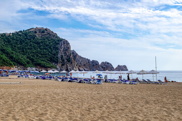 Alanya, Turkey - October 23, 2020: Umbrellas and sunbeds on the sandy Damlatas beach on the backdrop of a cliff with Alanya castle on top. Few people in a Turkish resort in the spring and autumn time