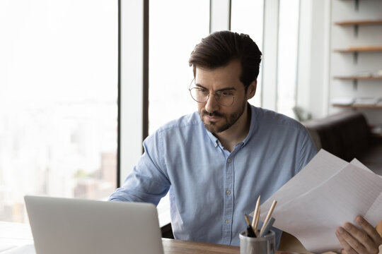 Smart Skilled Concentrated Young 30s Businessman Ceo Executive Manager In Glasses Analyzing Paper Documents, Working On Project Using Computer Software Application, Busy Workday In Modern Office Room.