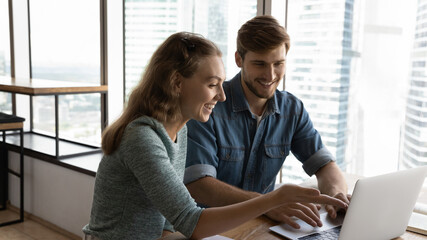 Happy female employee discussing online project ideas with male colleague, working together using computer applications, developing marketing or growth strategy together in modern office room.