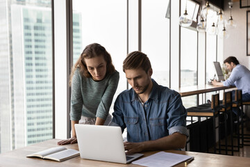 Concentrated young business lady helping male colleague with online computer project, working together at shared workplace. Focused smart workers analyzing online sales in modern office room.