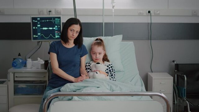 Portrait of worried mother and girl child looking into camera durinh illness therapy examination in hospital ward. Sad kid patient with nasal tube holding teedy bear resting in bed suffering infection