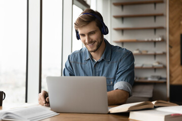 Smiling young handsome man in wireless headphones listening educational webinar lecture on computer, writing notes, improving professional knowledge, studying on online courses in modern home office.