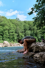 Young handsome Caucasian male traveler with dreadlocks and beard washes his feet in clean cold mountain river and sits on rock. Getting legs wet in icy river. Man is tourist.