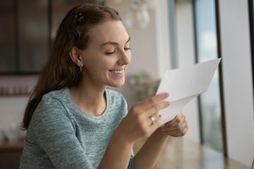 Smiling young woman reading paper correspondence letter with good news, feeling excited of getting...