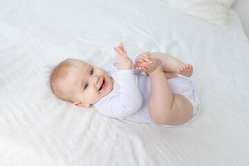 smiling baby girl six months old plays with her legs lying on her back on a white cotton bed in the bedroom of the house and laughs