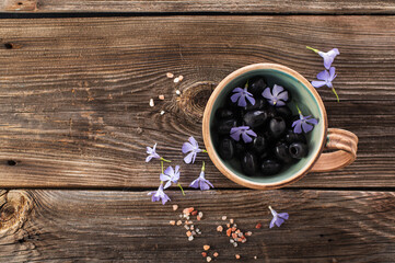 Obraz na płótnie Canvas black olives in a ceramic bowl on a wooden background, decorated with blue flowers and himalayan salt, top view, empty space for text
