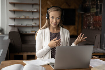 Happy beautiful young woman in wireless headphones improving knowledge, talking with teacher online, preparing for exams at home office. Smiling female student studying distantly on internet courses.