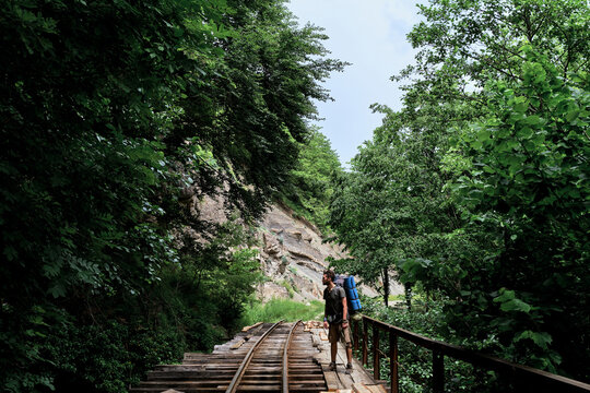 Handsome Caucasian man with beard and dreadlocks walks along old abandoned narrow gauge railway among dense summer forest. Tourist with large backpack stands on rails on wooden bridge.
