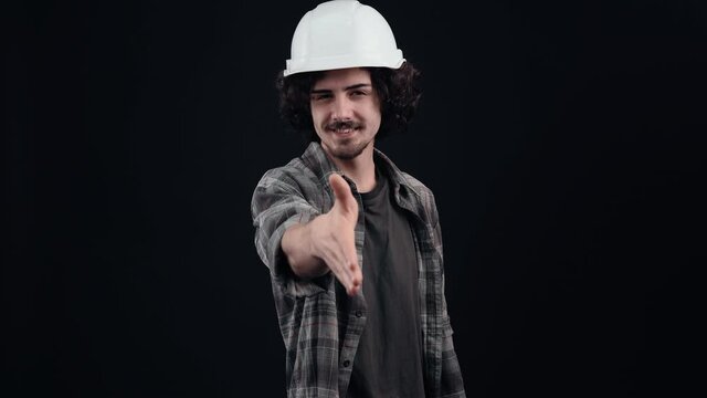 The novice, benevolent engineer, with a white helmet, extends his friendly hand to greet. Isolated on black background. The concept of life. People's emotions. 4k portrait