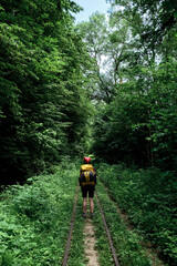 Woman tourist explores new places and trails. Caucasian female traveler is walking along railway with large yellow backpack among green summer forest. Rear view.