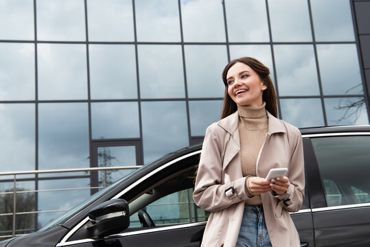 Cheerful Woman With Smartphone Looking Away While Standing Near Car