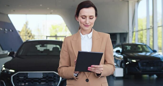 Portrait of confident smiling woman in formal wear using her tablet stands in front of cars in motor show looking into the camera. Professional saleswoman works in car dealership