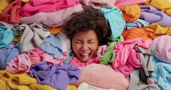 Emotional curly haired Afro American woman sticks out head through pile of multicolored unfolded clothes busy doing laundry at home screams loudly. Ethnic female cleans out wardrobe at home.