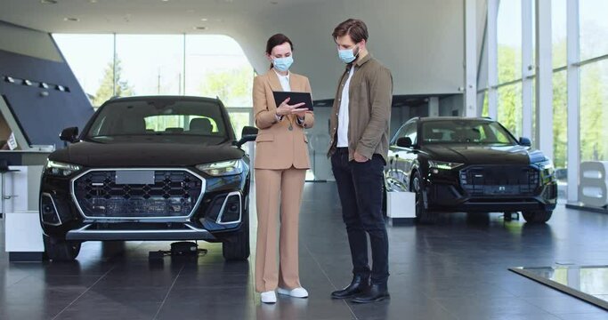 A male bearded client choosing car at dealership with the help of auto seller. Manager in auto dealership presents new cars using a tablet. Both in face masks.