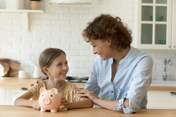 Happy small child girl sitting at table with affectionate young mother, holding piggybank in hands....