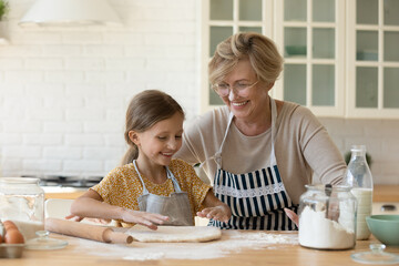 Happy diverse generations female family having fun, involved in preparing homemade pastry together in modern kitchen. Joyful old granny sharing culinary skills with cute little granddaughter.