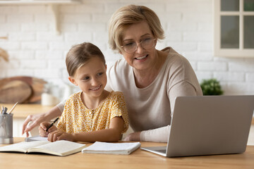 Caring middle aged retired grandmother in eyeglasses helping little 7s kid granddaughter with distant e-learning process, doing homework together or preparing for exams using computer at home.