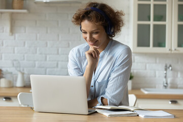 Happy young 30s attractive woman wearing wireless headphones, watching educational online seminar webinar, taking part in video conference or educational lecture, studying or working distantly at home