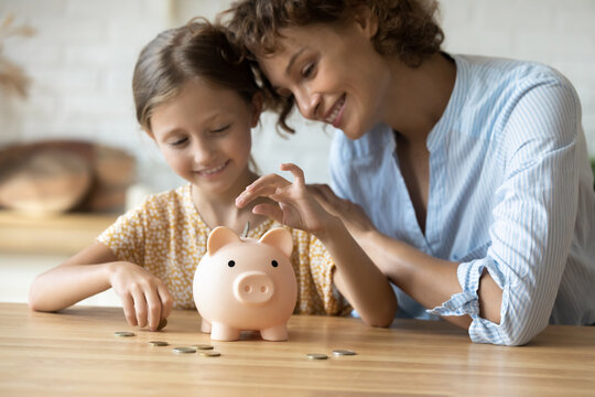Caring young mother teaching small preschool kid daughter saving money or planning future purchases, putting coins in small piggybank in modern kitchen, financial education for children concept.