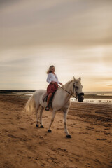 Fototapeta na wymiar Cute happy young woman on horseback in summer beach by sea. Rider female drives her horse in nature on evening sunset light background. Concept of outdoor riding, sports and recreation. Copy space